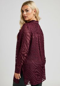 Burgunderfärgad genomskinlig blus med texturerat vågmönster, långa ärmar, krage och sidofickor för rörelsefrihet. Bakre veckdetalj synlig.