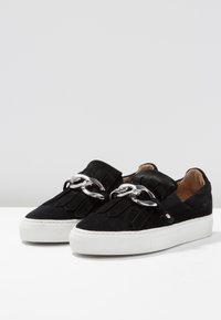 Chaussures sans lacets en daim noir avec un détail à franges et un accent de chaîne métallique. Semelle en caoutchouc blanche, bout rond, et design minimaliste.