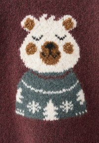 Pleteni pulover s licem medvjeda u krem bijeloj i smeđoj boji na tamnocrvenoj pozadini, sa zelenim ovratnikom s uzorkom koji prikazuje bijela stabla i pahulje.