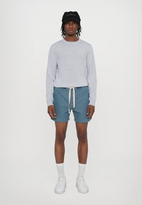 Chemise à manches longues gris clair, shorts en denim bleu avec cordon de serrage, chaussettes blanches et baskets blanches. Casquette noire en accent sur la tête.