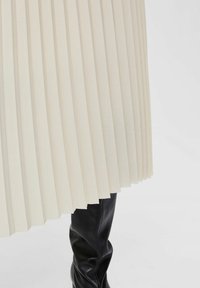 Gonna plissettata crema con una finitura testurizzata, abbinata a stivali in pelle neri al ginocchio, concentrandosi sulla struttura del tessuto e sul contrasto di colore.
