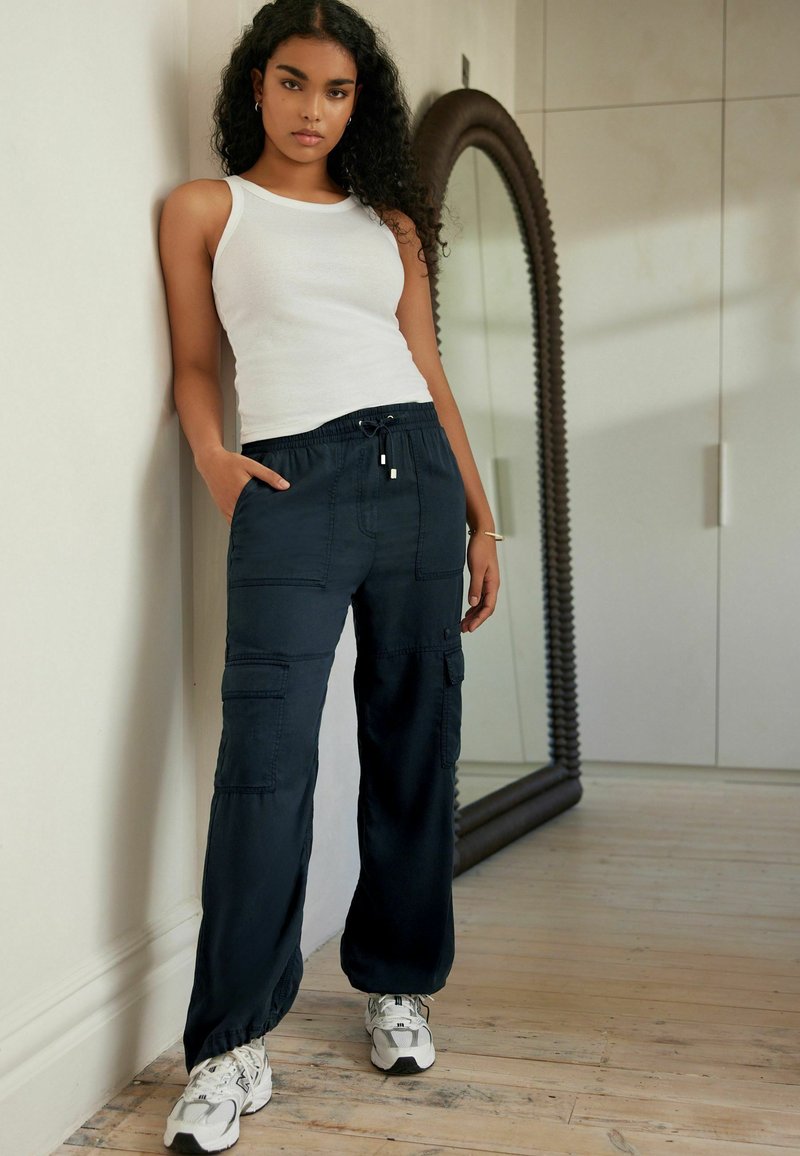 Pantaloni cargo navy con vita elastica, tasche laterali e vestibilità comoda, abbinati a una canotta bianca e sneakers grigi.