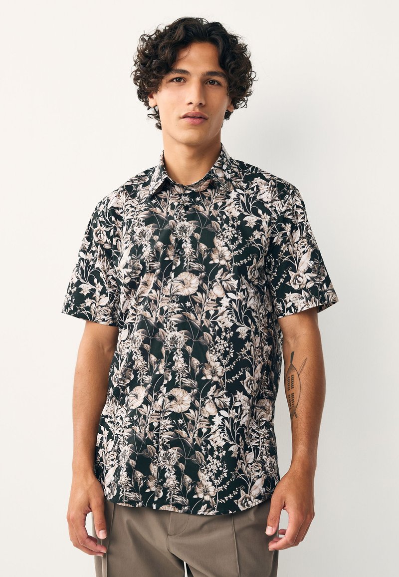 Jaunas vyras su garbanotais plaukais dėvi juodą trumpomis rankovėmis marškinėlį su kreminės spalvos gėlių raštu ir chaki spalvos kelnes, stovėdamas priešais baltą foną.