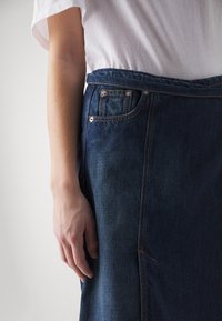 Džínová sukně s tmavým praním, se zvýšeným pasem, bočními kapsami a viditelným švem. Textura se jeví hladká s mírným leskem.