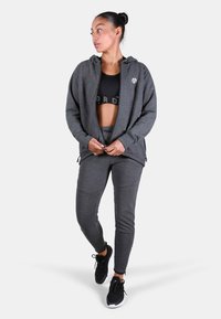 Šedá mikina na zips s kapucňou a bočnými vreckami, kombinovaná s čiernym crop topom a zodpovedajúcimi šedými nohavicami. Na hrudi má logo.