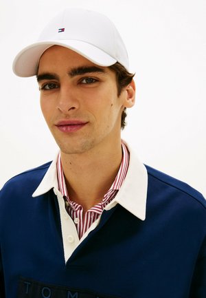 Jonge man met een witte pet, marineblauw shirt met een witte kraag, en daaronder een rood-wit gestreept overhemd met knopen, die licht glimlacht.