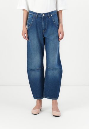 Jeans i denim med blå vask, vid ben-design, synlige sømmer og kne-patcher. Stylet med en hvit topp og rosa sko.
