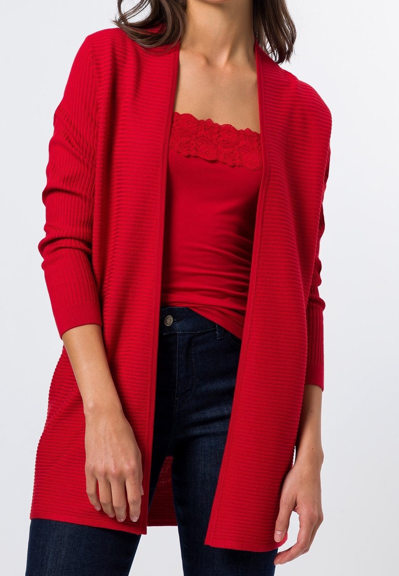 Femme portant un cardigan rouge à côtes ouvert sur le devant, par-dessus un caraco rouge orné de dentelle, associé à un jean bleu foncé.