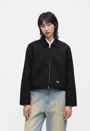 Jeune personne aux cheveux noirs mi-longs porte une veste zippée noire par-dessus une chemise blanche et un jean bleu clair délavé.