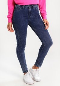 Smala, högmidjade jeans i mörkblå denim med en urtvättad textur, kombinerat med vita sneakers och en rosa sweatshirt.
