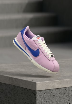 Heled lilla Nike tossud sinise swooshiga ja valgete paeltega, mis hõljuvad betoonist ääre kohal, taustal trepid.