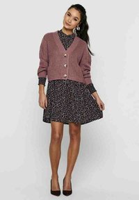 Stofroze gebreide cardigan met een V-hals, knoopsluiting en geribbelde textuur, gedragen over een zwarte bloemenjurk met lange mouwen.