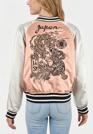 Vrouw draagt een satijnen bomberjack met een roze achterpaneel met zwarte borduursels van een draak en tijger en de tekst "Japan", gecombineerd met lichtblauwe jeans.