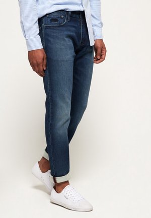 Homme portant un jean bleu foncé retroussé, une chemise rayée bleu clair et des baskets blanches, debout devant un fond uni.