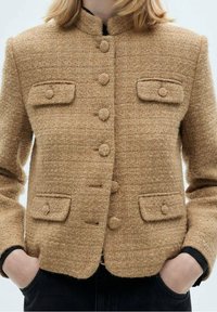 Veste beige texturée à boutons avec quatre poches à rabat, portée sur des manches noires, mains dans les poches, sur un fond clair uni.