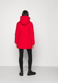 Parka roja aislada con capucha, que presenta una cintura ajustada, mangas largas y un dobladillo recto. Se lleva con leggings negros y botas de tobillo.