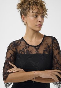 Mujer con cabello rizado en un vestido de encaje negro, brazos cruzados, usando pendientes de aro dorados y una pulsera delicada, mirando hacia un lado contra un fondo blanco.