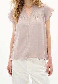 Blusa di un leggero lavanda con maniche a farfalla, scollo a V e pieghe frontali. Realizzata in un tessuto morbido, abbinata a pantaloni chiari.