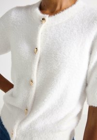 Hvid fluffy cardigan med korte ærmer, prydet med gyldne knapper foran, struktureret overflade og en rund halsudskæring.