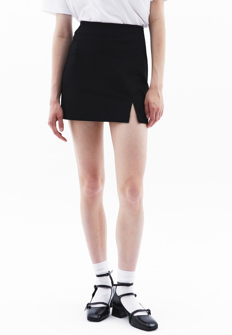 Zwarte mini rok met een splits aan de voorkant, gecombineerd met witte sokken en zwarte Mary Jane schoenen. Geschikt voor een gladde stof en een op maat gemaakte pasvorm.