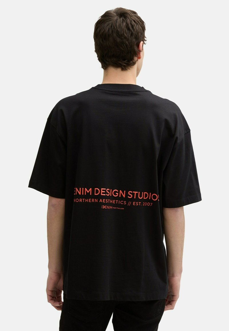 Black oversized T-shirt made of cotton with short sleeves. Features graphic text in red on the back, stating "DENIM DESIGN STUDIO."
