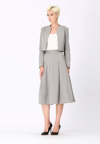 Veste grise courte sur un haut blanc avec une jupe grise pleine, faite d'un tissu lisse, au design épuré et dotée de détails plissés, associée à des talons noirs.