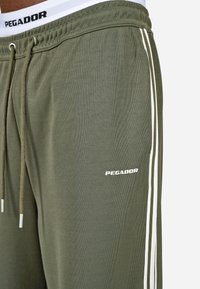 Olive green sweatpants with white side stripes, drawstring waistband, and a logo reading "PEGADOR" in white on the left thigh.