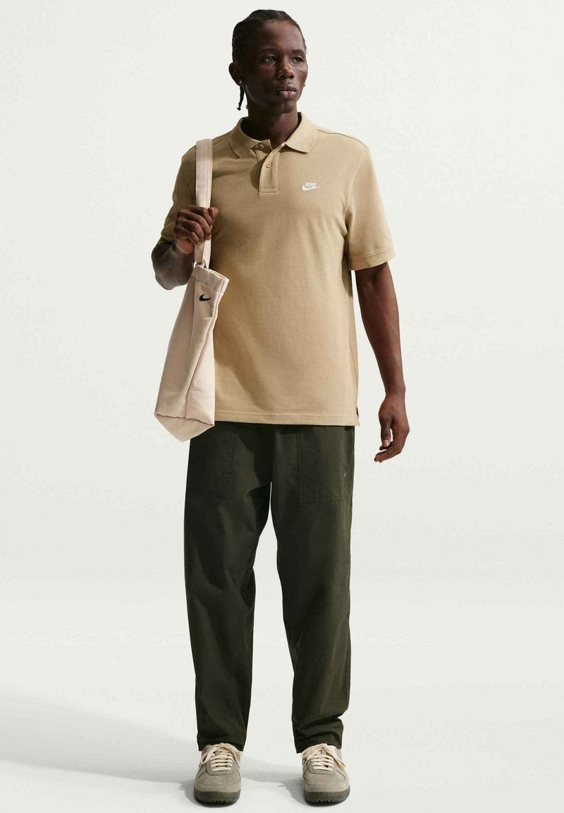 Polo beige avec un petit logo blanc, pantalon cargo vert olive, chaussures beiges, portant un tote bag beige clair avec un logo.