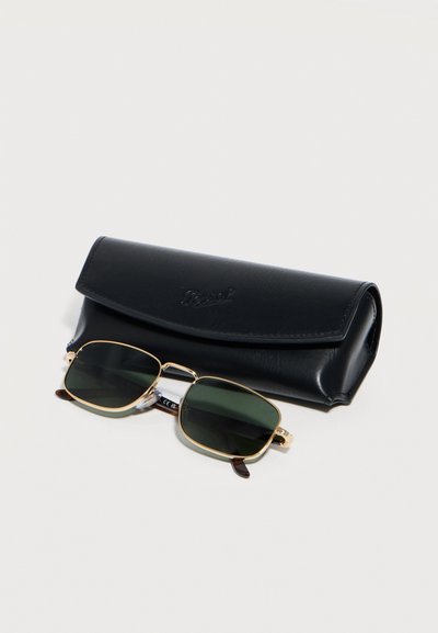 Ορθογώνια γυαλιά ηλίου με χρυστούς μεταλλικούς σκελετούς και σκουρόχρωμους φακούς, τοποθετημένα μπροστά από μια μαύρη θήκη προστασίας από δέρμα.