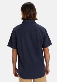 Camisa de manga corta azul marino con cuello, con yugo plano en la parte trasera y una textura suave, que presenta un dobladillo curvado y sin cierres visibles.