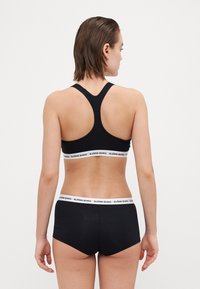 Μαύρο αθλητικό σουτιέν με σχέδιο racerback και λευκή επωνυμία στη ζώνη. Συνδυάζεται με μαύρα boyshorts που διαθέτουν ταιριαστή ζώνη με λογότυπο.