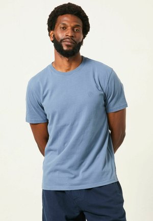 Mand med skæg og afro står med hænderne bag ryggen iført en lyseblå kortærmet T-shirt og mørkeblå bukser mod en ensfarvet baggrund.