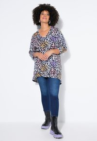 Frau mit lockigem Haar, die ein mehrfarbiges Leopardenmuster-Top, blaue Jeans und schwarze Stiefel mit lila Sohlen trägt, vor einem weißen Hintergrund stehend.