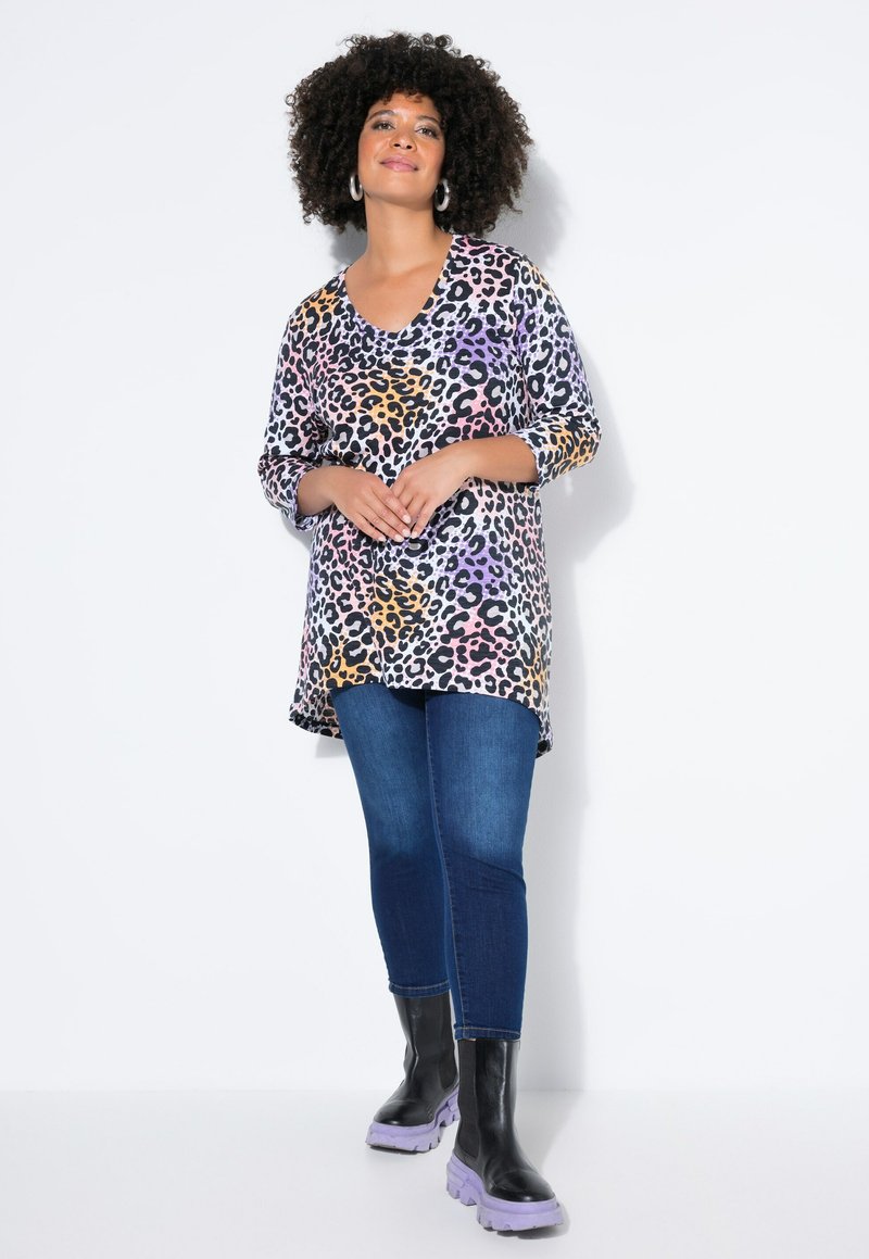 Frau mit lockigem Haar, die ein mehrfarbiges Leopardenmuster-Top, blaue Jeans und schwarze Stiefel mit lila Sohlen trägt, vor einem weißen Hintergrund stehend.