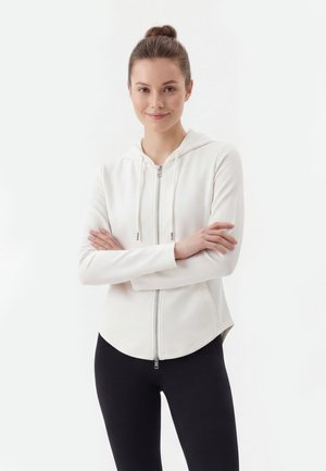 Młoda kobieta z włosami związanymi w kok, ubrana w białą bluzę z zamkiem i czarne legginsy, stoi z założonymi ramionami i delikatnie się uśmiecha.