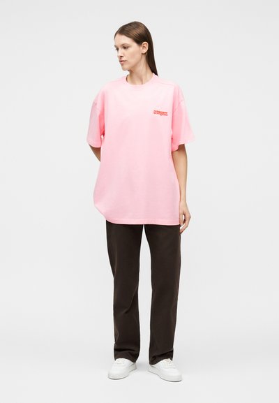 Γυναίκα που στέκεται, φοράει φαρδιά ροζ μπλούζα με κόκκινο κείμενο, σκούρο ίσιο παντελόνι και λευκά αθλητικά παπούτσια, στρέφοντας το βλέμμα της προς τα αριστερά πάνω σε ένα απλό φωτεινό φόντο.