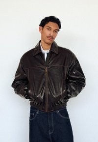 Young man with curly hair wearing a dark brown leather jacket over a white shirt and dark jeans, standing against a plain white wall.