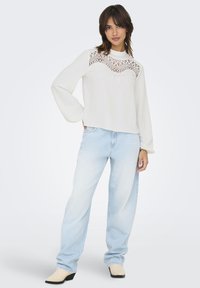White sweater with intricate lace detailing at the neckline, paired with light blue straight-leg jeans and cream-colored heeled boots.