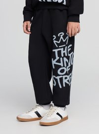 Czarne spodnie dresowe z napisem „THE KING OF STREET” w dużym, jasnoniebieskim kolorze na lewej nogawce. Do tego białe tenisówki z czarnymi paskami.