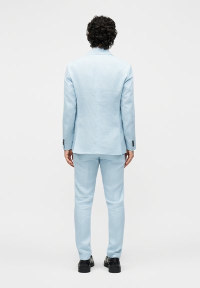 Άνδρας με σγουρά σκούρα μαλλιά στέκεται με την πλάτη γυρισμένη, φοράει ένα ανοιχτό μπλε κοστούμι και μαύρα επίσημα παπούτσια σε μια απλή λευκή φόντο.