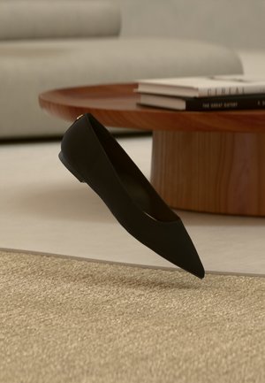 Pantof cu talpă joasă, vârf ascuțit, de culoare neagră, plutind deasupra unei covor bej, cu o masă de cafea din lemn și cărți stivuite în fundal.