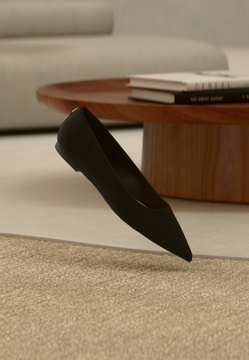 Μαύρο επίπεδο παπούτσι με μυτερή μύτη που αιωρείται πάνω από μπεζ χαλί με ξύλινο τραπεζάκι καφέ και στοιβαγμένα βιβλία στο παρασκήνιο.