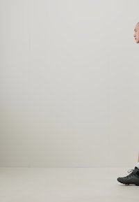 Chaussures de sport noires avec une pointe arrondie et une surface texturée, portées avec des chaussettes blanches. Personne marchant sur un fond uni de couleur claire.
