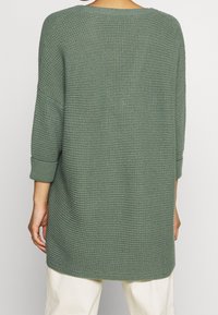 Maglione verde oversize a maglia con un motivo testurizzato, spalle scese e polsini a coste. L'orlo arriva fino a metà coscia.
