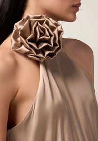 Beige satiini mekko, jossa on roikkuva muotoilu ja suuri teksturoitu ruusukoriste olkapäällä, jossa on pehmeitä, laskostettuja terälehtiä.