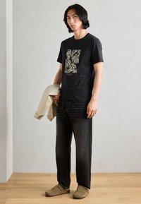 T-shirt nera con stampa grafica, abbinata a jeans scuri e giacca chiara. Il modello indossa scarpe marroni slip-on. Sfondo semplice.