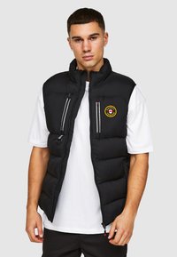 Zwarte gewatteerde vest met hoge kraag, ritssluiting aan de voorkant en twee ritssluitingen op de borst. Bevat een cirkelvormig geel logo aan de linkerkant.