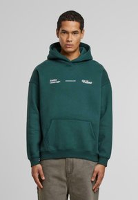 Sudadera de algodón verde con bolsillo canguro, con texto impreso en blanco y un dobladillo acanalado. Textura suave y corte relajado.