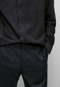 Dunkel gestreiftes Hemd mit Knöpfen, kombiniert mit anthrazitfarbenen Hosen. Das Hemd hat eine glatte Textur; die Hosen sind leicht strukturiert und mit Taschen versehen.