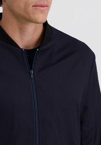 Man in een donkerblauw zip-up jack met zwarte kraag en een zwart shirt eronder, weergegeven van de nek tot halverwege de borst tegen een effen achtergrond.